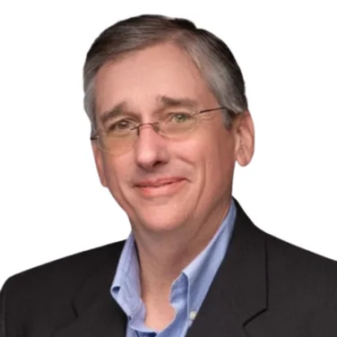 A middle-aged man wearing glasses, a blue button-up shirt, and a black blazer poses against a white background.