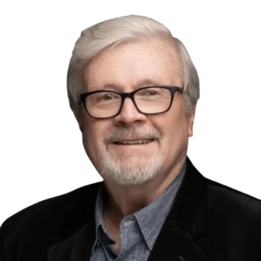 Smiling older man with white hair, glasses, and a short beard, wearing a dark blazer over a blue button-up shirt, against a white background.