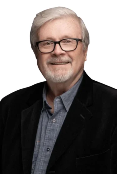 Smiling older man with white hair, glasses, and a short beard, wearing a dark blazer over a blue button-up shirt, against a white background.