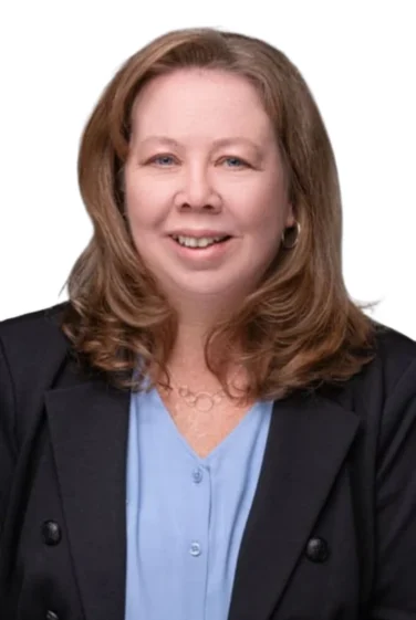 Woman with shoulder-length light brown hair wearing a black blazer and light blue blouse, smiling at the camera against a plain white background.