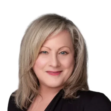 A woman with shoulder-length blonde hair, wearing a black blazer over a red top and a long silver necklace, poses against a plain white background.