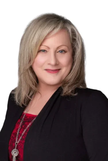 A woman with shoulder-length blonde hair, wearing a black blazer over a red top and a long silver necklace, poses against a plain white background.