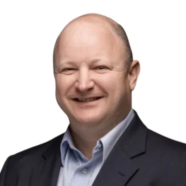 A bald man wearing a dark suit jacket and a light blue collared shirt, smiling at the camera against a plain white background.