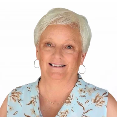 Older woman with short white hair wearing a sleeveless floral blouse and hoop earrings, smiling at the camera against a plain white background.