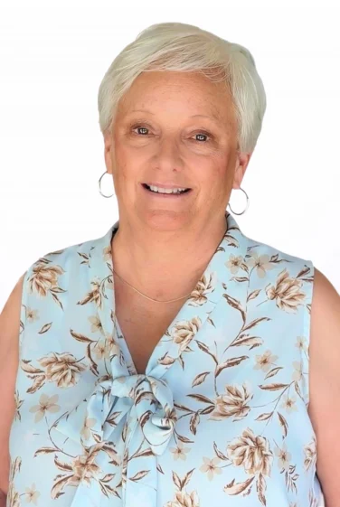 Older woman with short white hair wearing a sleeveless floral blouse and hoop earrings, smiling at the camera against a plain white background.