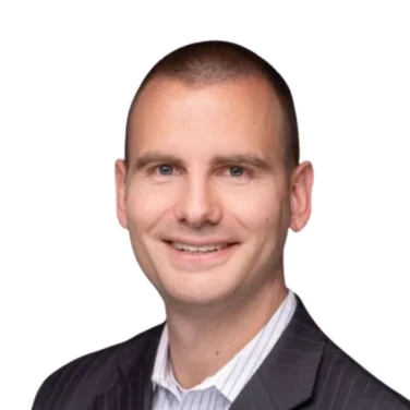 Man in a dark pinstripe suit jacket and light striped shirt, smiling, with a shaved head, photographed against a plain white background.
