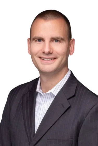 Man in a dark pinstripe suit jacket and light striped shirt, smiling, with a shaved head, photographed against a plain white background.