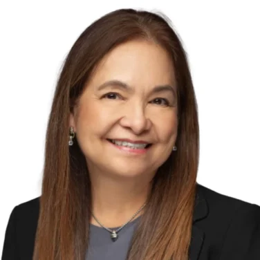 Woman with long straight brown hair, wearing a dark blazer over a gray top, smiling at the camera, against a white background.