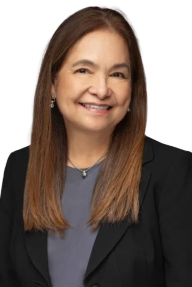 Woman with long straight brown hair, wearing a dark blazer over a gray top, smiling at the camera, against a white background.