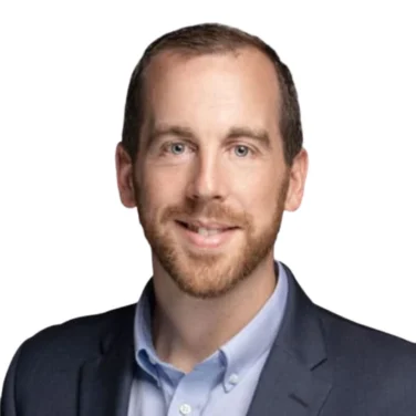 Man with short brown hair and beard wearing a navy blazer and light blue collared shirt, smiling, against a plain white background.