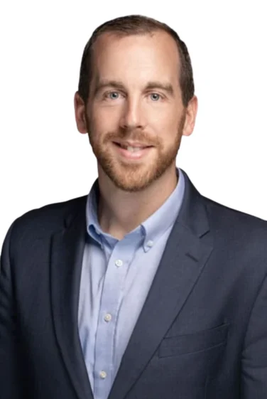 Man with short brown hair and beard wearing a navy blazer and light blue collared shirt, smiling, against a plain white background.