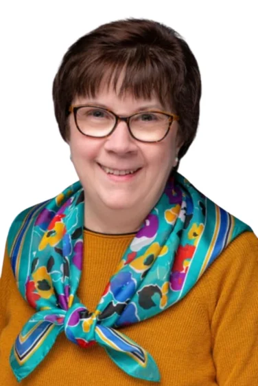 Smiling woman with short brown hair, glasses, and a colorful floral scarf over a mustard yellow top, against a white background.