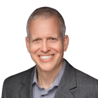 Smiling adult man with short gray hair, wearing a gray blazer over a collared shirt, against a plain white background.