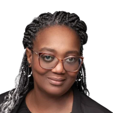 A woman with braided hair, glasses, and a black top and jacket, poses against a plain white background.