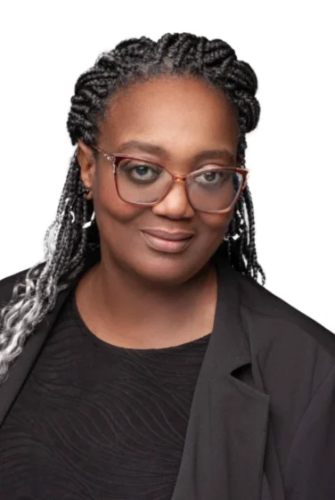 A woman with braided hair, glasses, and a black top and jacket, poses against a plain white background.