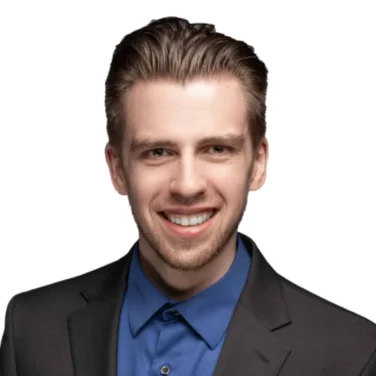 Smiling young man with short brown hair wearing a dark blazer and blue dress shirt, photographed against a white background.