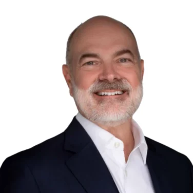 Bald man with a gray beard wearing a dark suit jacket and white shirt, smiling, against a plain white background.