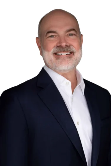 Bald man with a gray beard wearing a dark suit jacket and white shirt, smiling, against a plain white background.