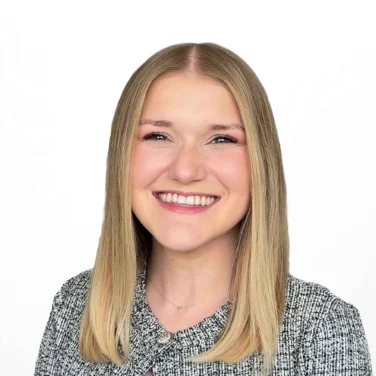 Bree Gore, a woman with straight, shoulder-length blonde hair, smiles while wearing a textured gray jacket over a black top, standing against a plain white background.