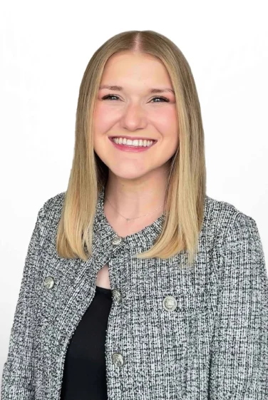 Bree Gore, a woman with straight, shoulder-length blonde hair, smiles while wearing a textured gray jacket over a black top, standing against a plain white background.