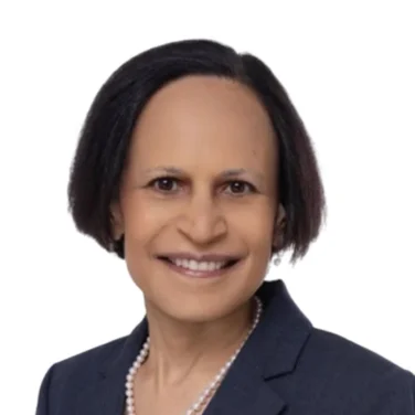 Smiling person with short dark hair wearing a navy blazer, light blouse, and pearl necklace, posed against a plain white background.