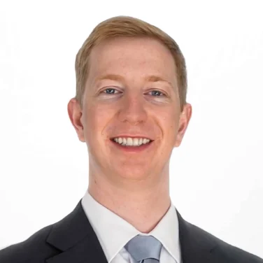 A man wearing a black suit, white shirt, and light blue tie is smiling at the camera against a plain white background.