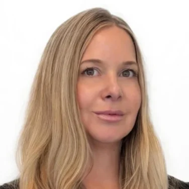 Woman with long, wavy blonde hair wearing a black top, posing in front of a plain white background.