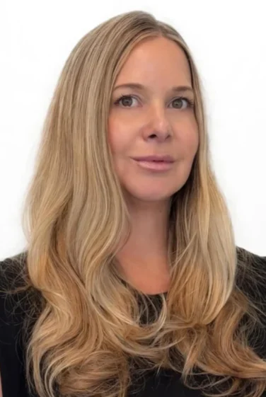 Woman with long, wavy blonde hair wearing a black top, posing in front of a plain white background.