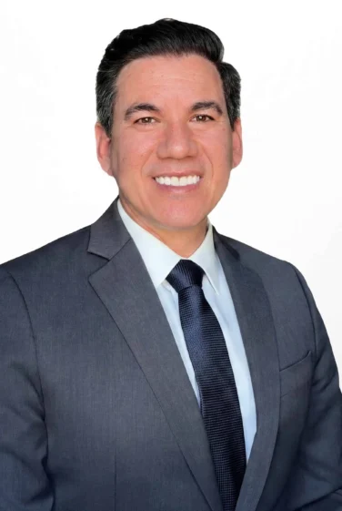 A man in a gray suit and dark tie is smiling at the camera against a plain white background.