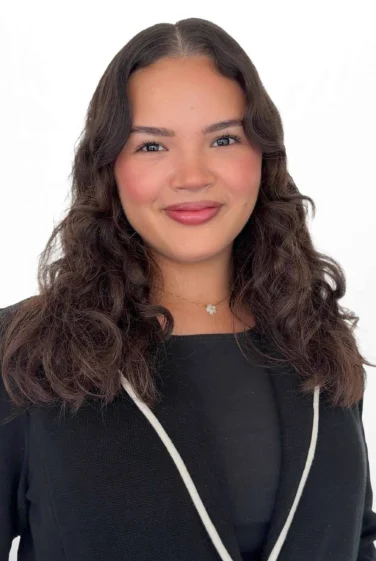 A woman with wavy dark hair and a black top smiles at the camera against a white background.