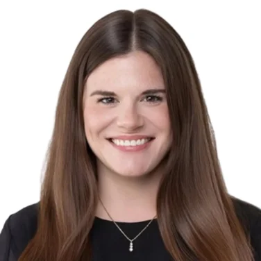 A woman with long brown hair, wearing a black top and a necklace, smiles while facing the camera against a white background.
