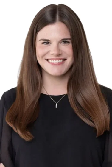 A woman with long brown hair, wearing a black top and a necklace, smiles while facing the camera against a white background.