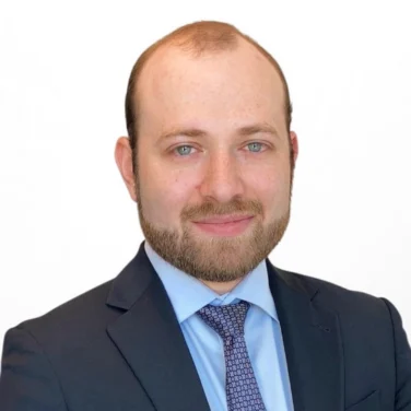 A man with a beard in a dark suit, light blue shirt, and patterned tie stands against a plain white background, looking at the camera.