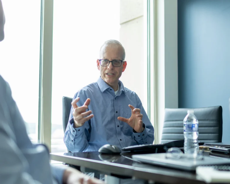 An older man wearing glasses gestures while speaking in a modern office meeting room with another person, water bottle, and laptop on the table.