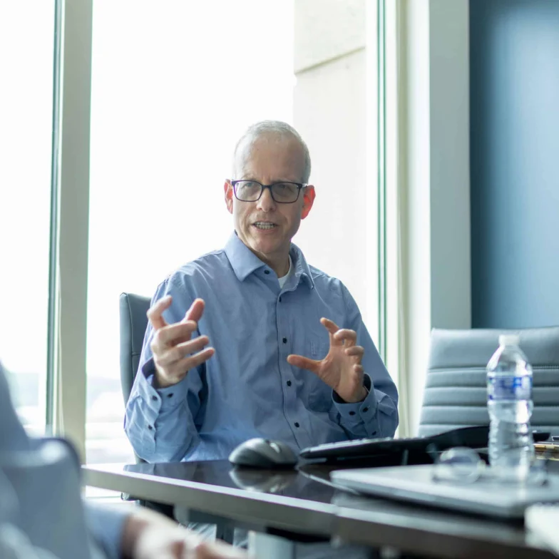 Older man in glasses sits at a conference table, gesturing with his hands while speaking to someone off camera. Office setting with a laptop, water bottle, and large window in background.