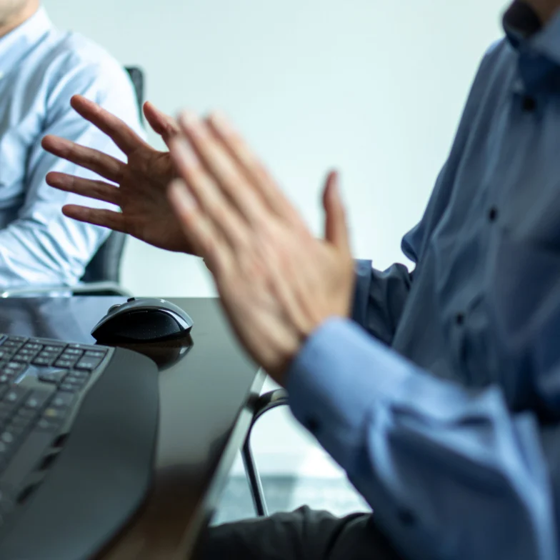 Two people in blue shirts sit at a desk with a keyboard and mouse; one is gesturing with hands while talking, the other listens.