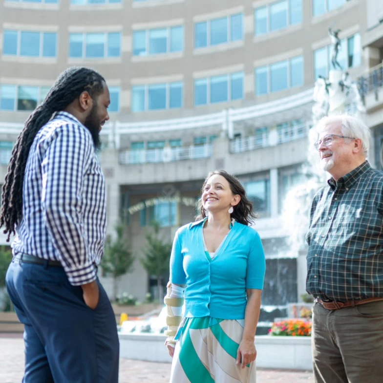 Three people stand in a courtyard having a conversation, with a fountain and tall building in the background.