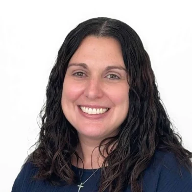 Smiling woman with long, wavy dark hair wearing a navy blue top and a necklace with a cross pendant, posed against a plain white background.