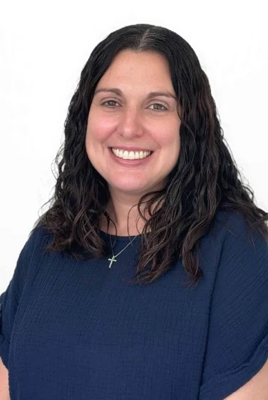 Smiling woman with long, wavy dark hair wearing a navy blue top and a necklace with a cross pendant, posed against a plain white background.