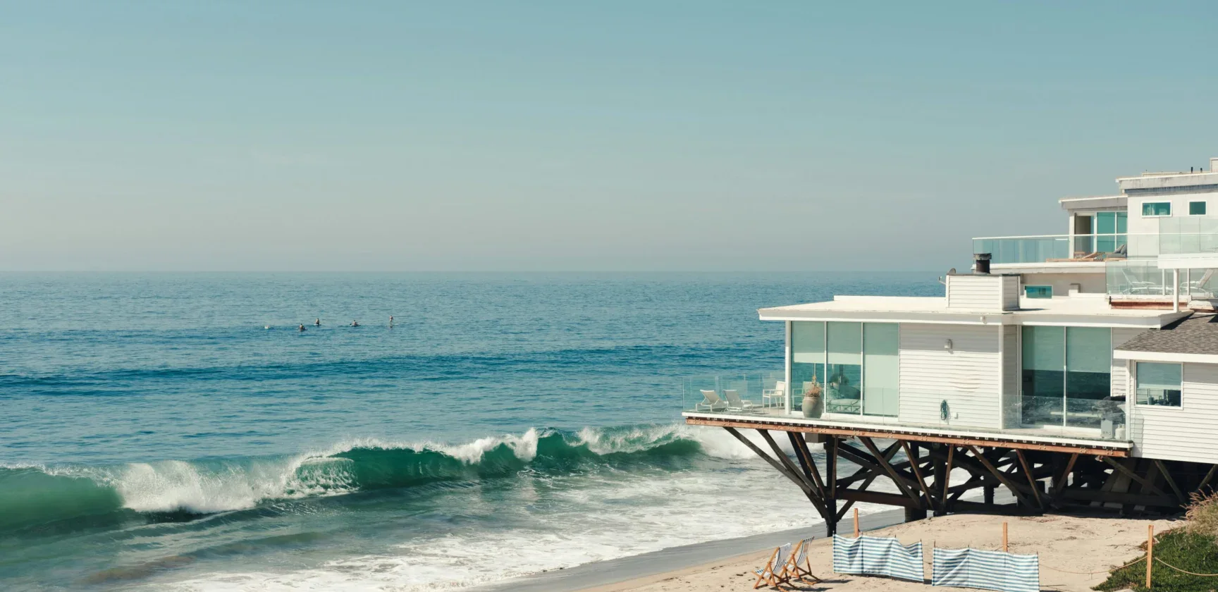 Modern house on stilts overlooks a sandy beach with waves and a few people in the ocean under a clear sky. Beach chairs and umbrellas sit on the sand in the foreground.
