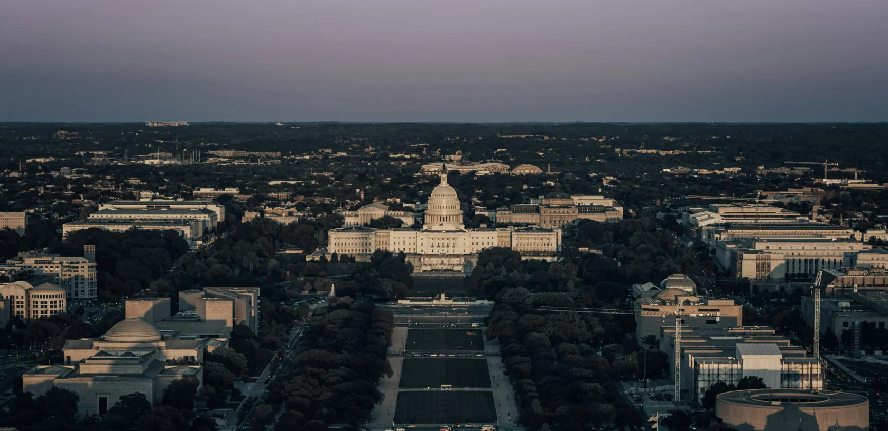 Aerial view of the United States Capitol building in Washington, D.C., with surrounding government buildings and tree-lined streets at dusk.