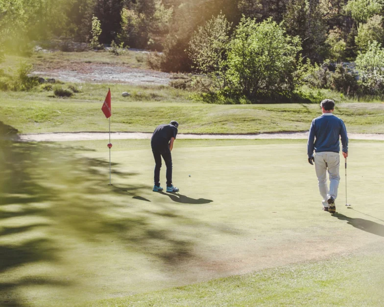 Two people are on a golf green; one is putting toward the hole while the other stands nearby holding a golf club. Trees and grass surround the area.