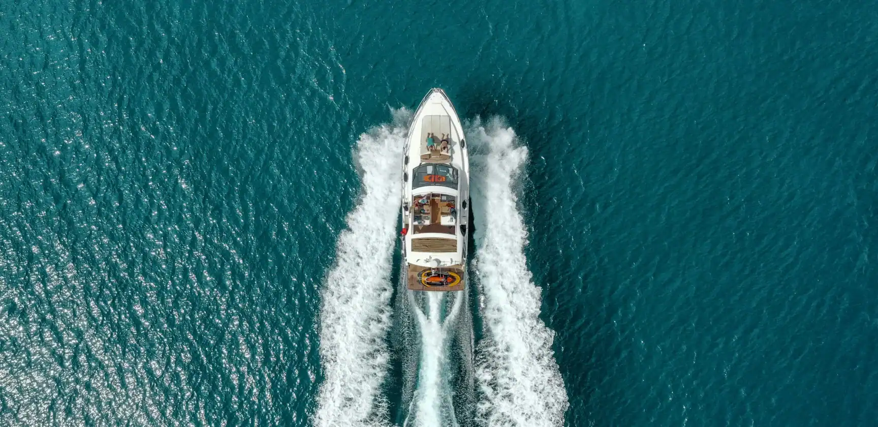 A white motorboat speeds through clear blue water, creating waves behind it. The image is taken from above, showing the boat’s wake on the surface.
