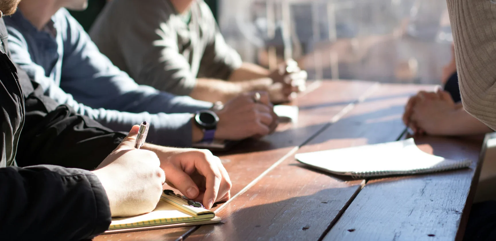 Four people sit around a wooden table in sunlight, taking notes and discussing, with notebooks and pens visible.