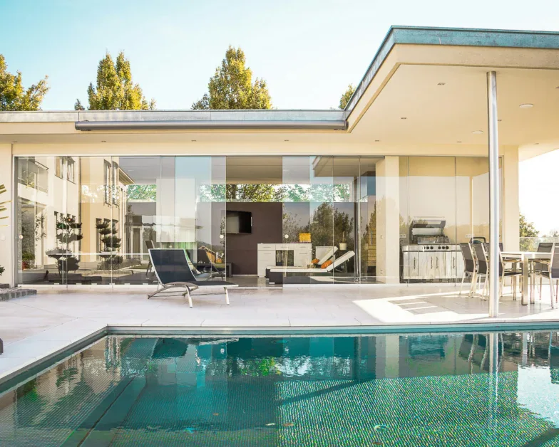Modern house with large glass windows, outdoor lounge chairs, a dining area, grill, and swimming pool on a sunny day. Trees are visible in the background.
