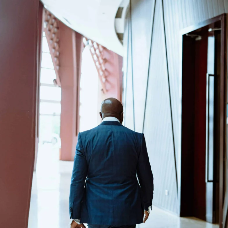 A person in a blue suit walks down a modern hallway with large windows and geometric wall designs, holding something in their left hand.