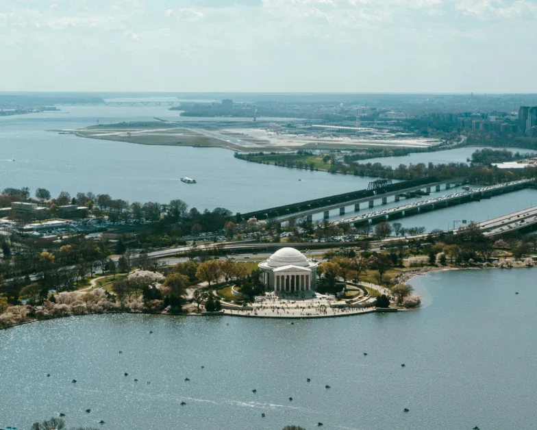 Aerial view of the Jefferson Memorial surrounded by water and cherry blossom trees, with bridges and cityscape visible in the background under a partly cloudy sky.