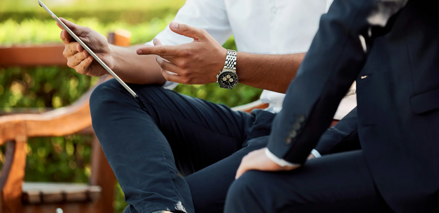 Two people sit outdoors on a bench, discussing something on a tablet. Both are dressed in business attire. Only their torsos and hands are visible.