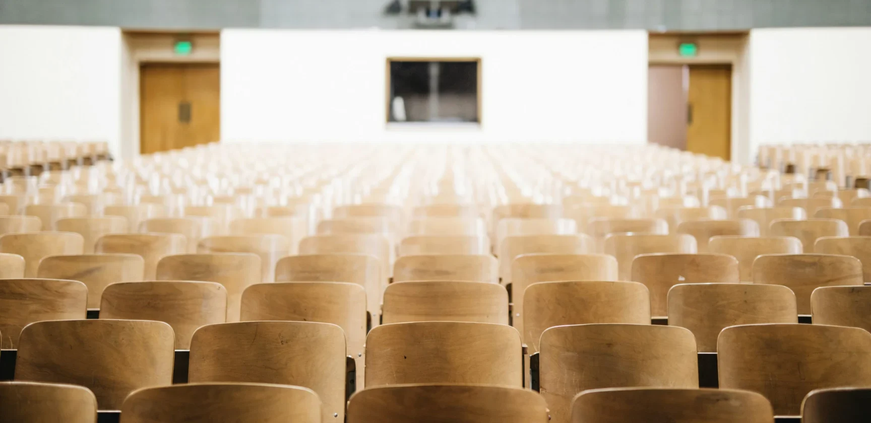 Empty rows of wooden seats in a large lecture hall facing a white wall with a clock and two closed doors.