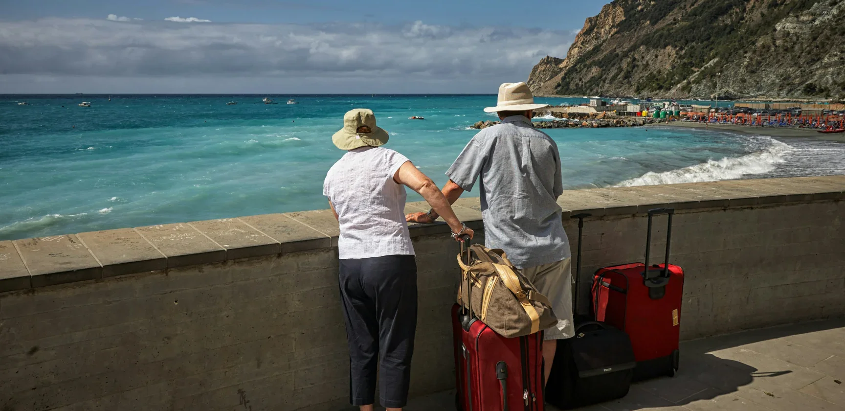 Two people wearing hats stand by a seaside railing with red suitcases, looking at the ocean and distant beach under a partly cloudy sky.